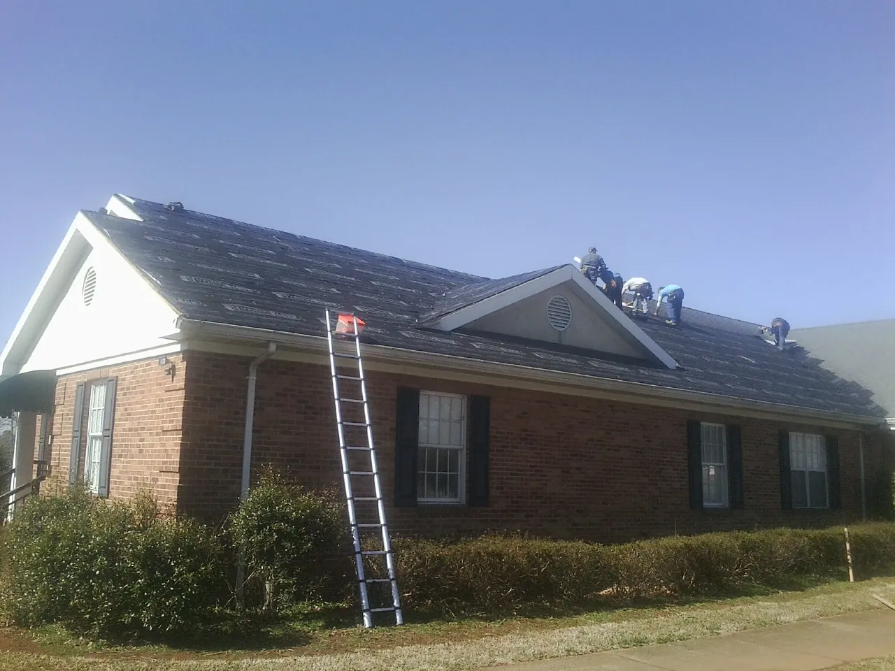 Crew tearing shingles off a brick ranch roof with a ladder set — Emergency Roof Repair work by our crew in Bushnell