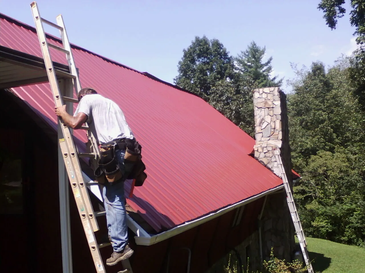 Roofer on a ladder beside a red metal roof and stone chimney — Soffit & Fascia Repair by Roofing Brothers in Bushnell